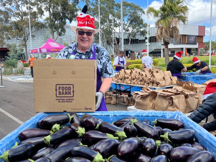 Foodbank Staff Member Steve at a market wearing a Christmas hat, holding a hamper in front of a crate of eggplants